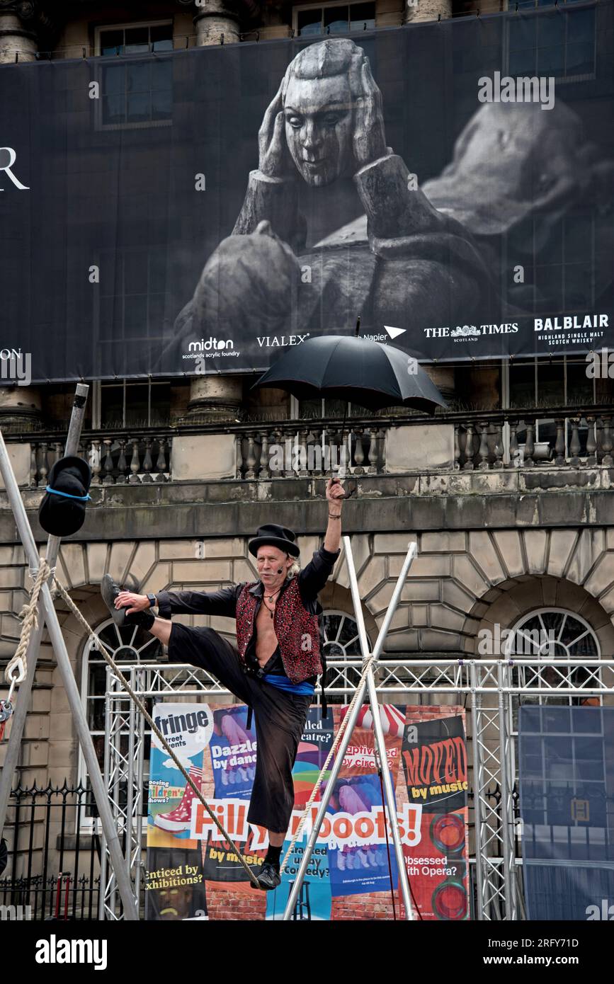 Street performer walking a tightrope during the Edinburgh Fringe Festival, in the background a banner advertises a David Eustace exhibition. Stock Photo