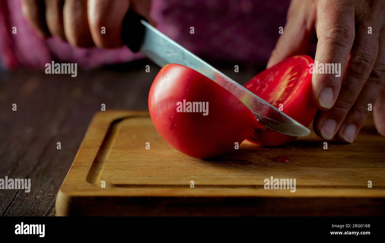 A woman cuts a red tomato with a knife on a wooden plank Stock Photo