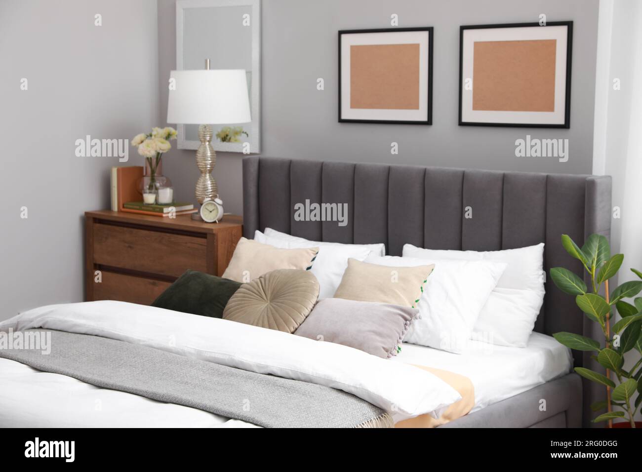 Comfortable bed with cushions, lamp and different decor on wooden chest of drawers in room. Stylish interior Stock Photo