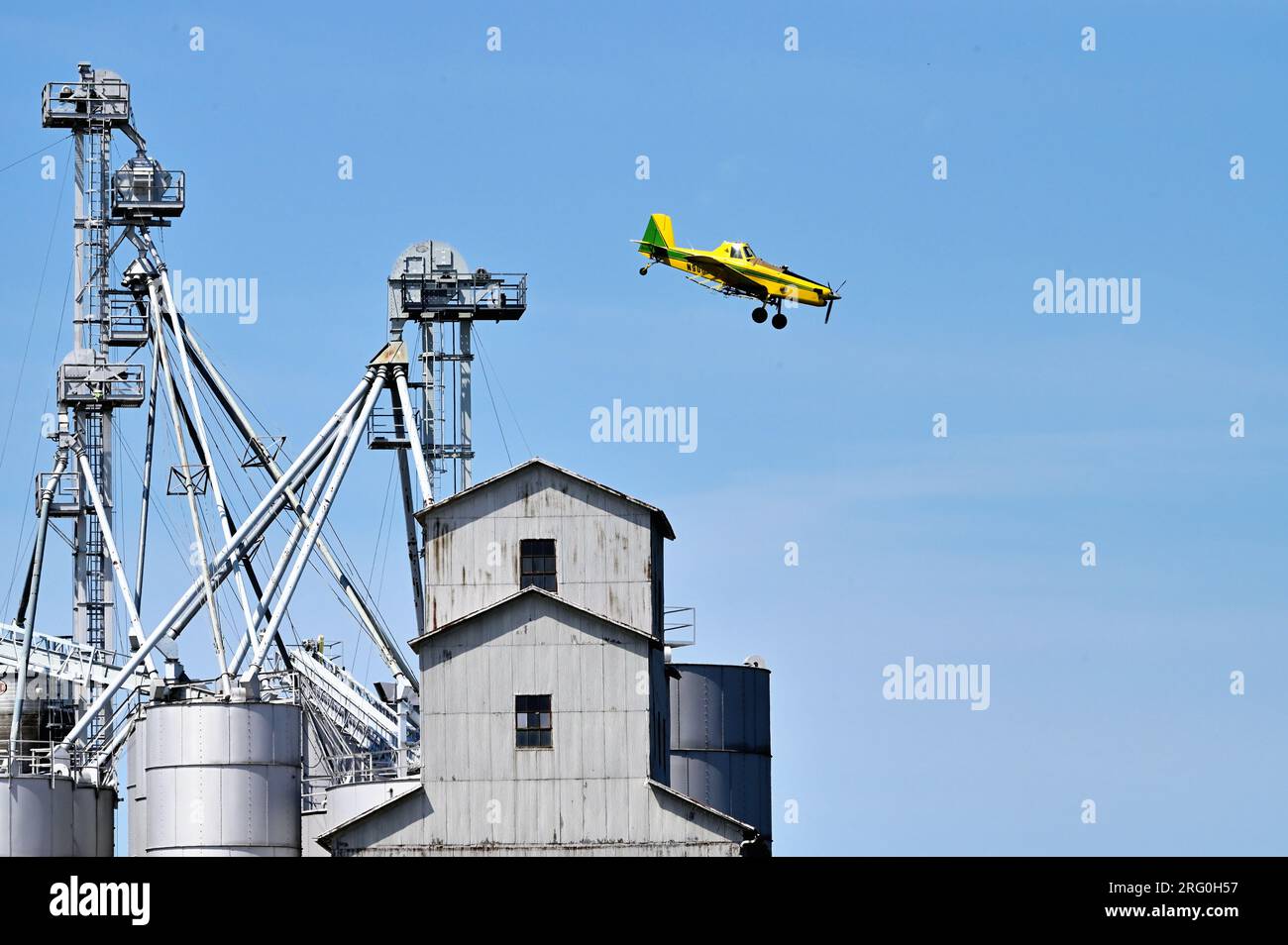 Earlville, Illinois, USA. A crop dusting airplane in a dive approaching a field of crops while passing very near a large grain elevator. Stock Photo