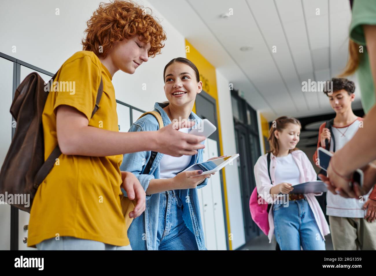 back to school, classmates using gadgets in school hallway, redhead boy using smartphone near girl Stock Photo