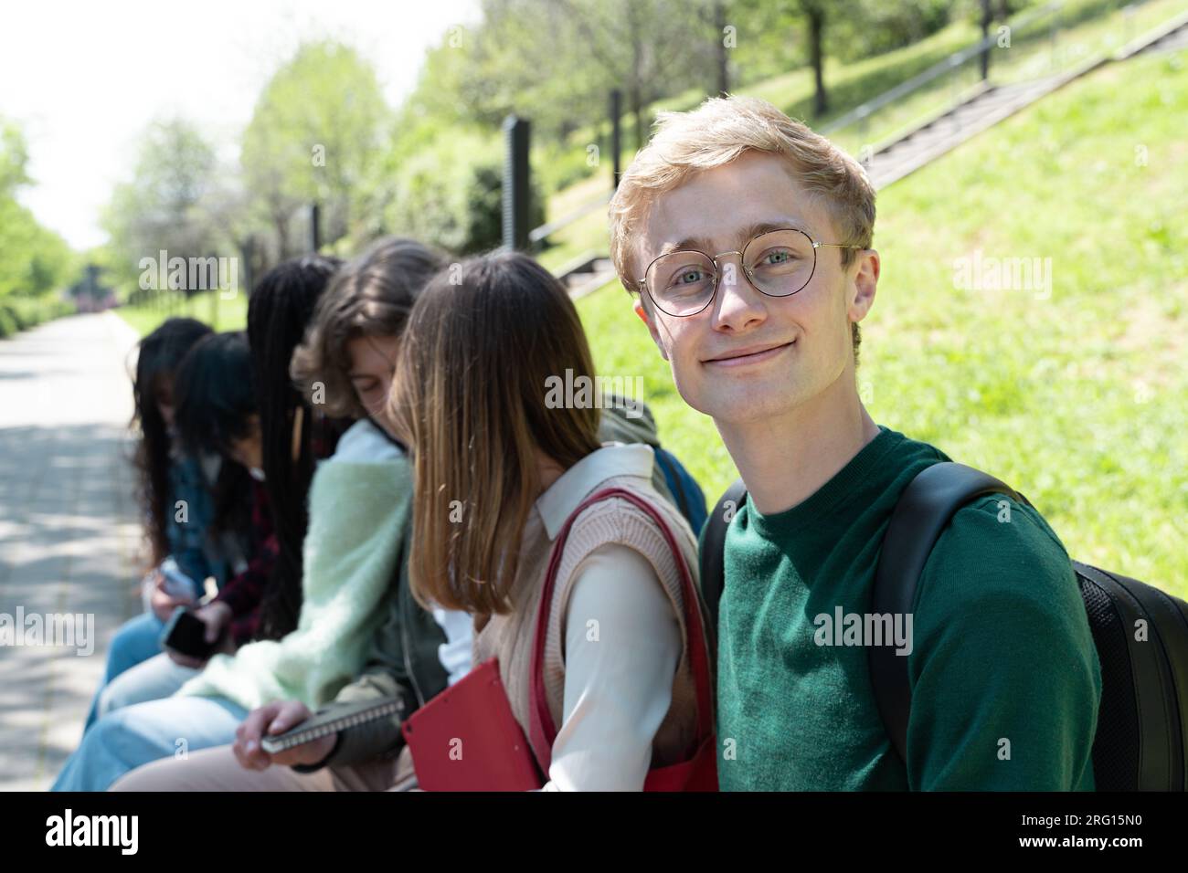 A group of multiracial teen students sits in a park. While friends interact with each other, a British boy with glasses, blond hair, and freckles smil Stock Photo