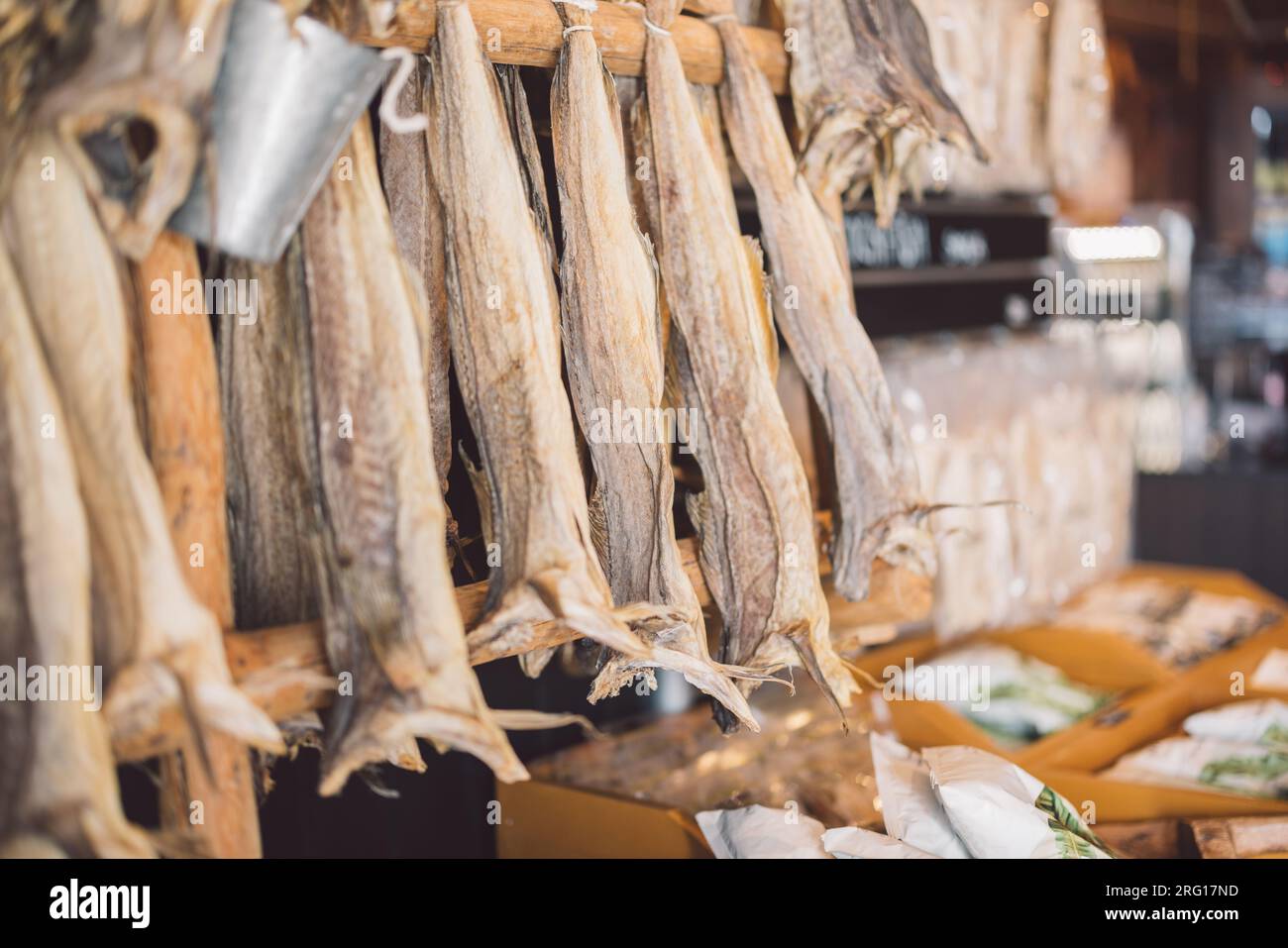 Cod dried fish stock hanging from wooden log in blurred shop with assorted fresh marine species in trays near weighing machine in sunny daylight in La Stock Photo