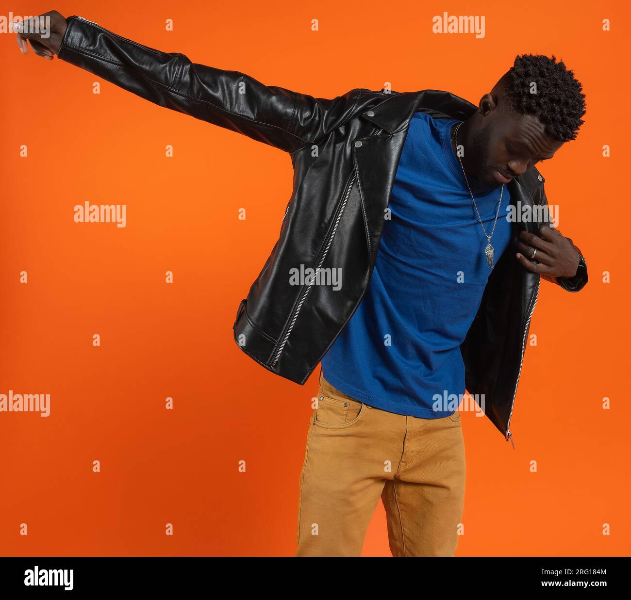 Young African American male with dark curly hair looking down while standing stretching arm and wearing black leather jacket against orange background Stock Photo