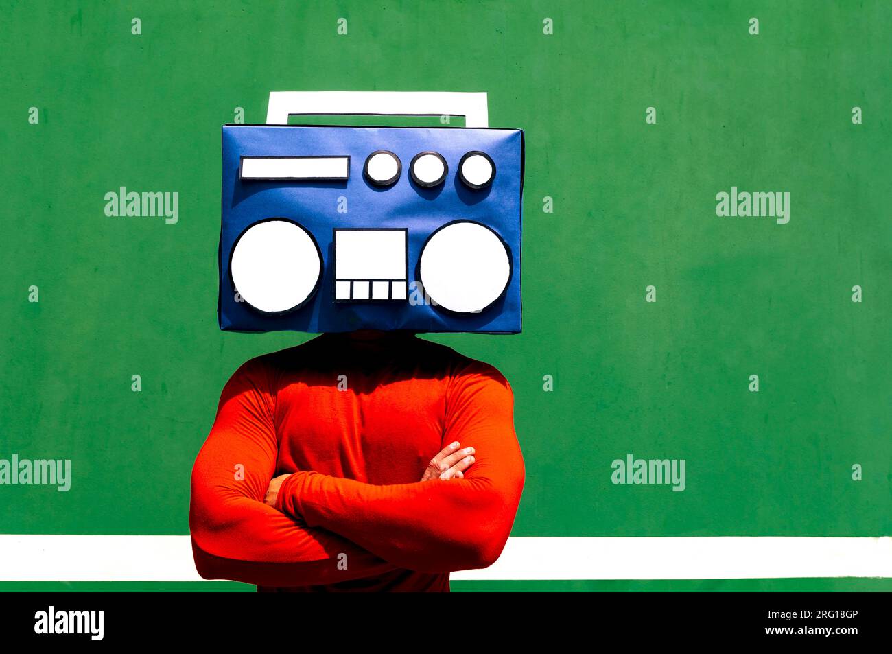 Unrecognizable person in red casual clothes standing arms crossed with blue boombox covering face against green background Stock Photo