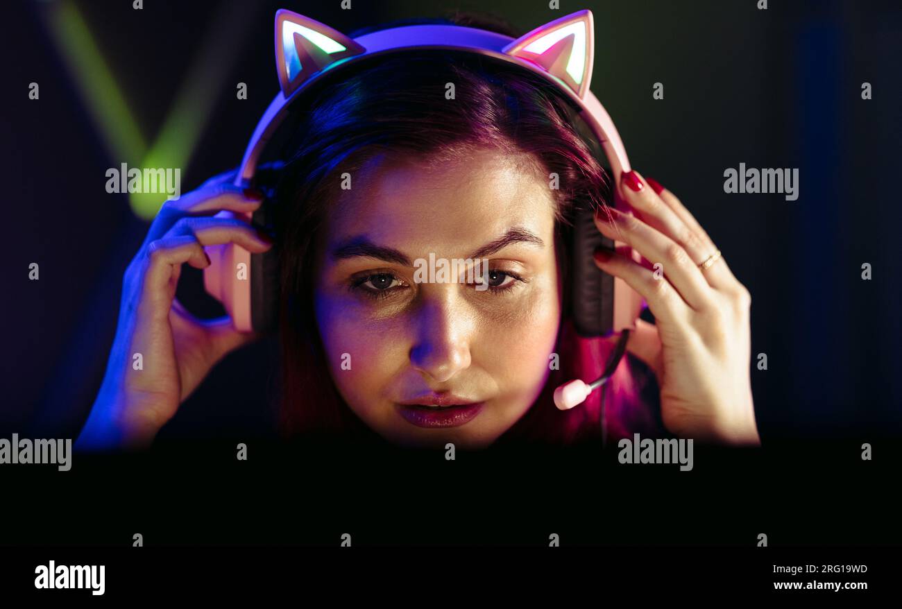 Woman adjusting her headset while looking at her gaming computer with concentration. Dedicated female gamer staying up all night playing video games o Stock Photo
