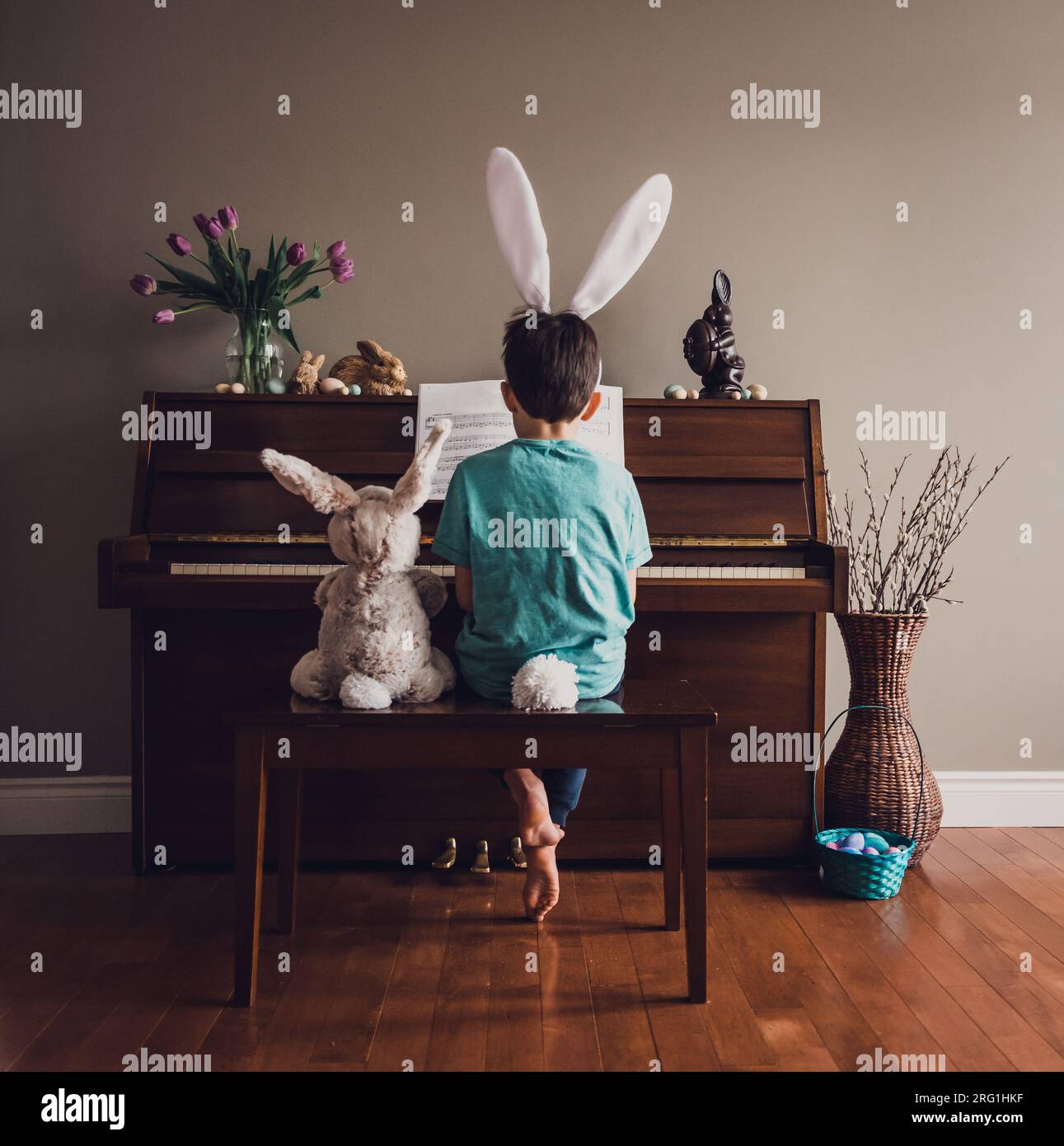 Boy wearing bunny ears playing piano with stuffed rabbit beside him. Stock Photo