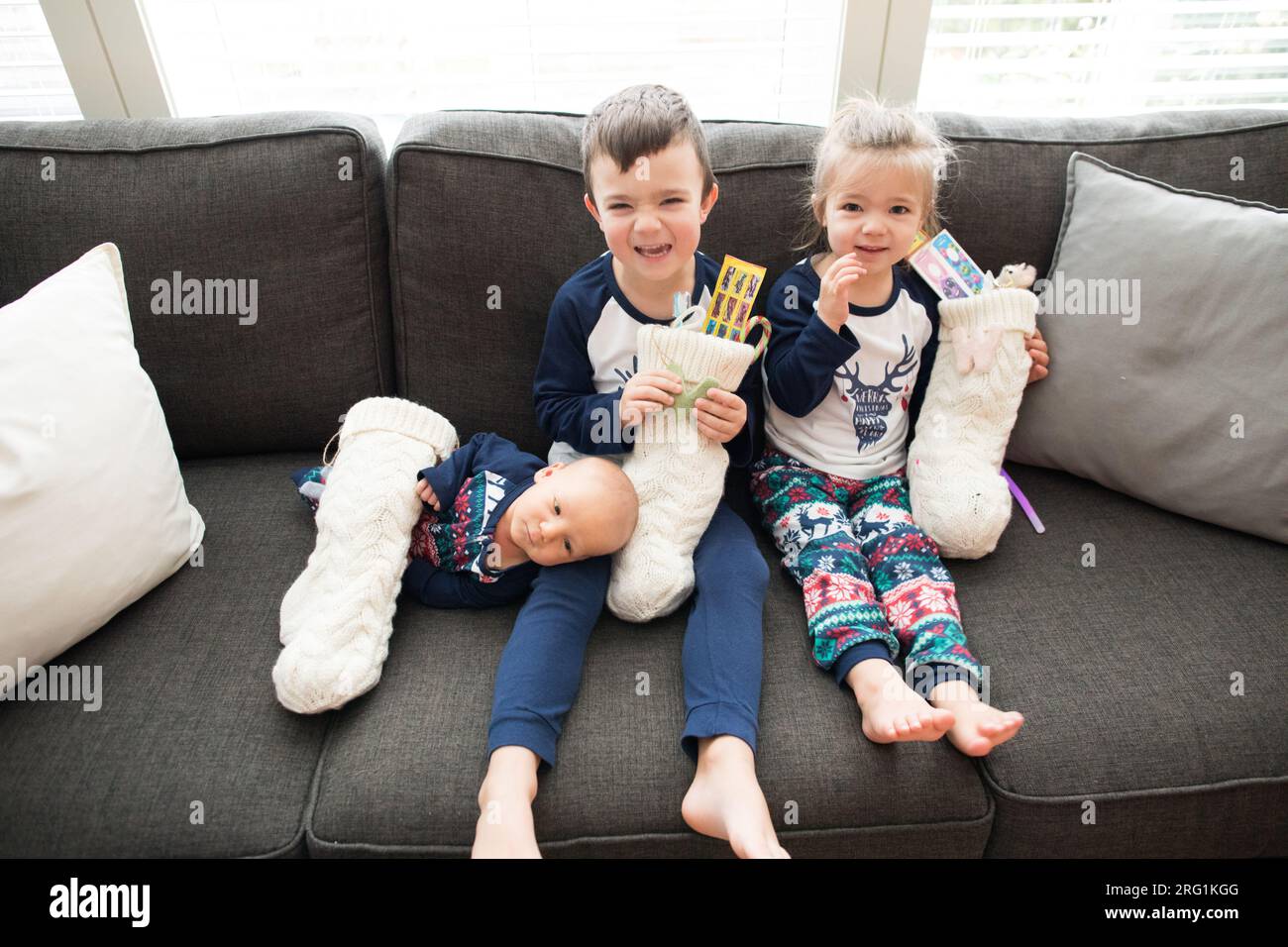 Three kids sit on couch holding their Christmas stockings Stock Photo