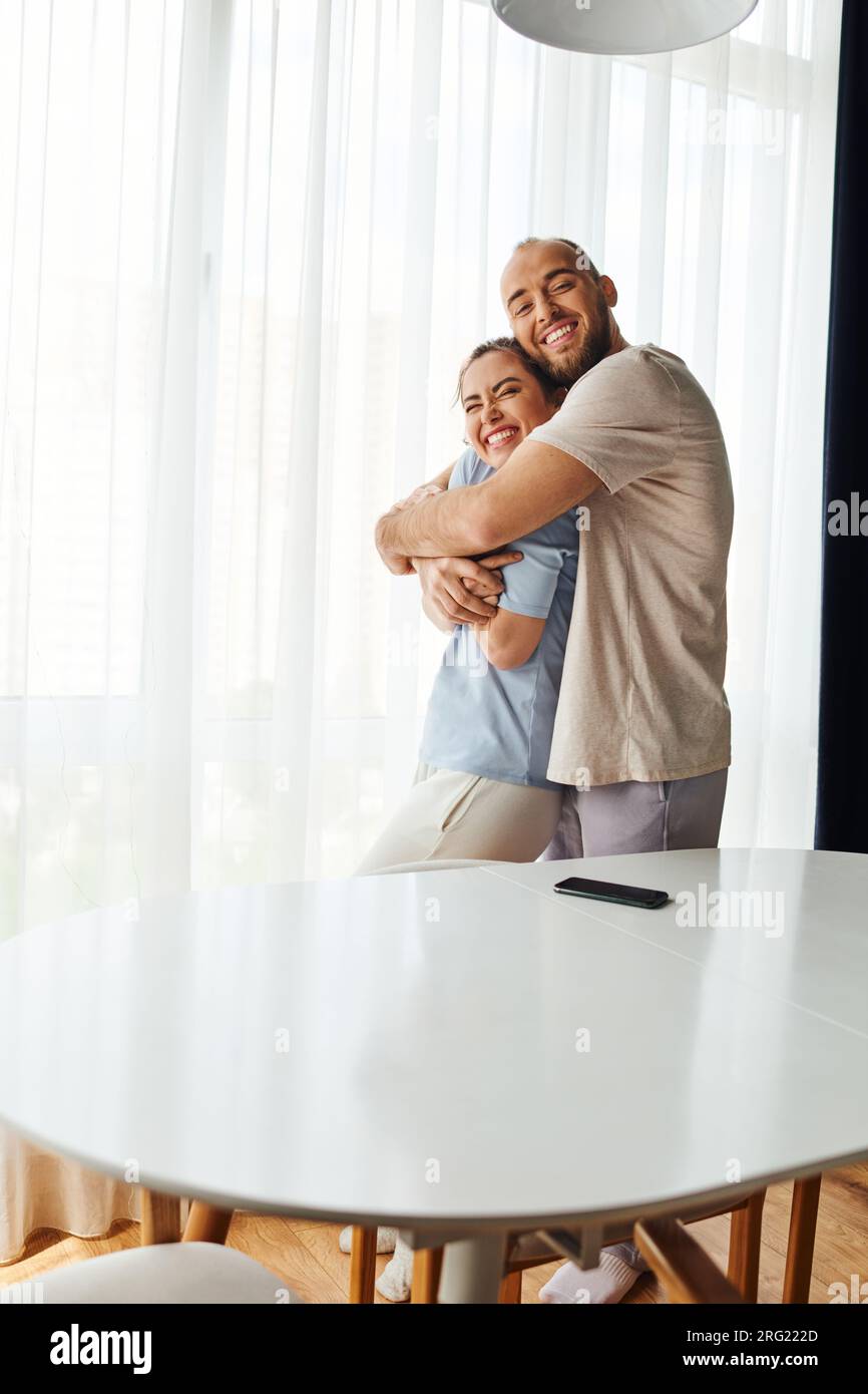 Smiling bearded man in homewear hugging girlfriend near smartphone on table at home Stock Photo
