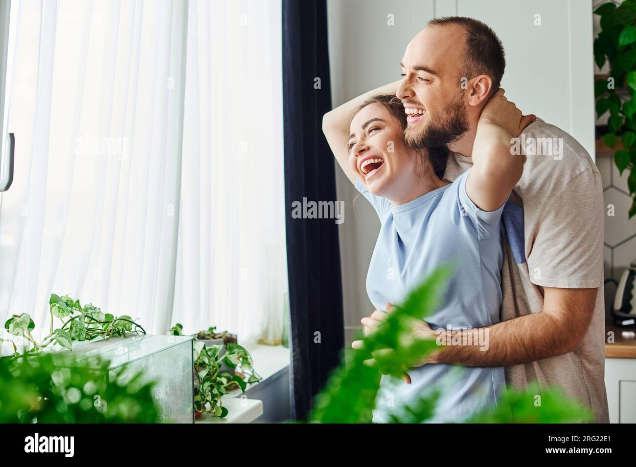 Joyful and relaxed couple in homewear hugging while standing near window at home Stock Photo