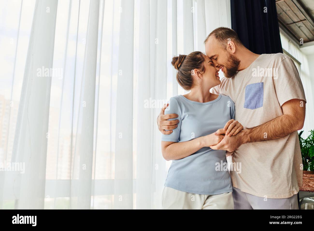 Side view of smiling couple in homewear holding hands and hugging while standing at home Stock Photo