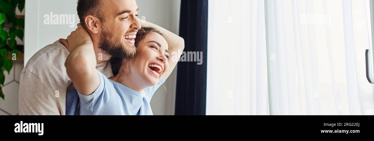 Positive woman hugging boyfriend in homewear while standing together near window at home, banner Stock Photo
