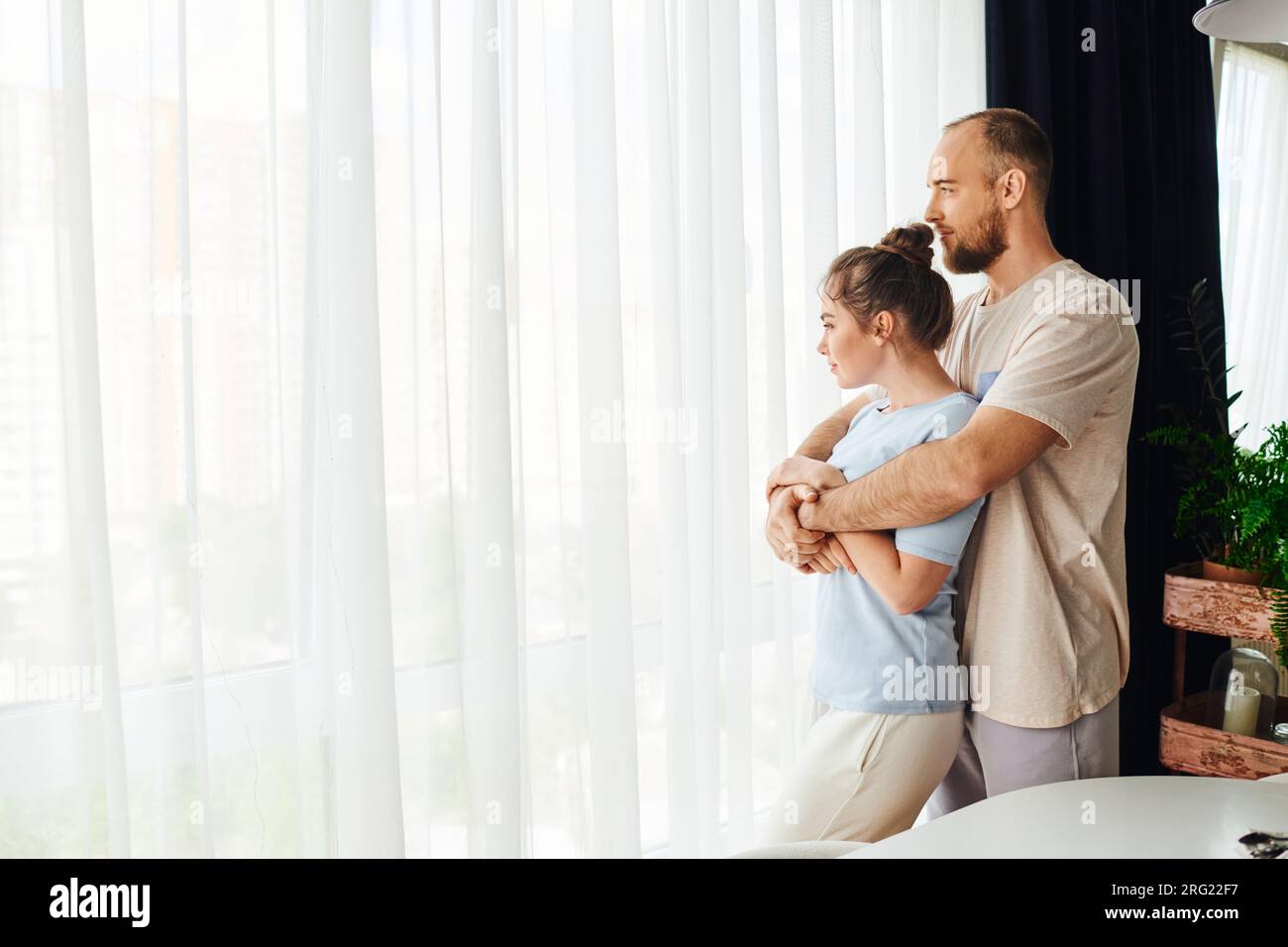 Side view of bearded man in homewear hugging girlfriend while standing near window at home Stock Photo