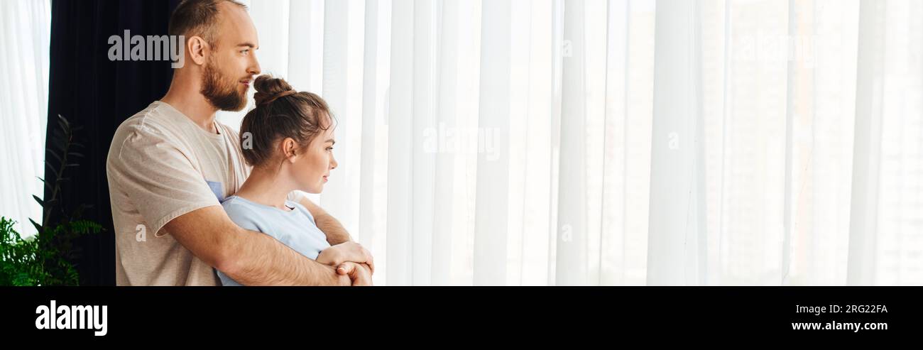 Side view of bearded man in loungewear hugging girlfriend near curtain at home, banner Stock Photo