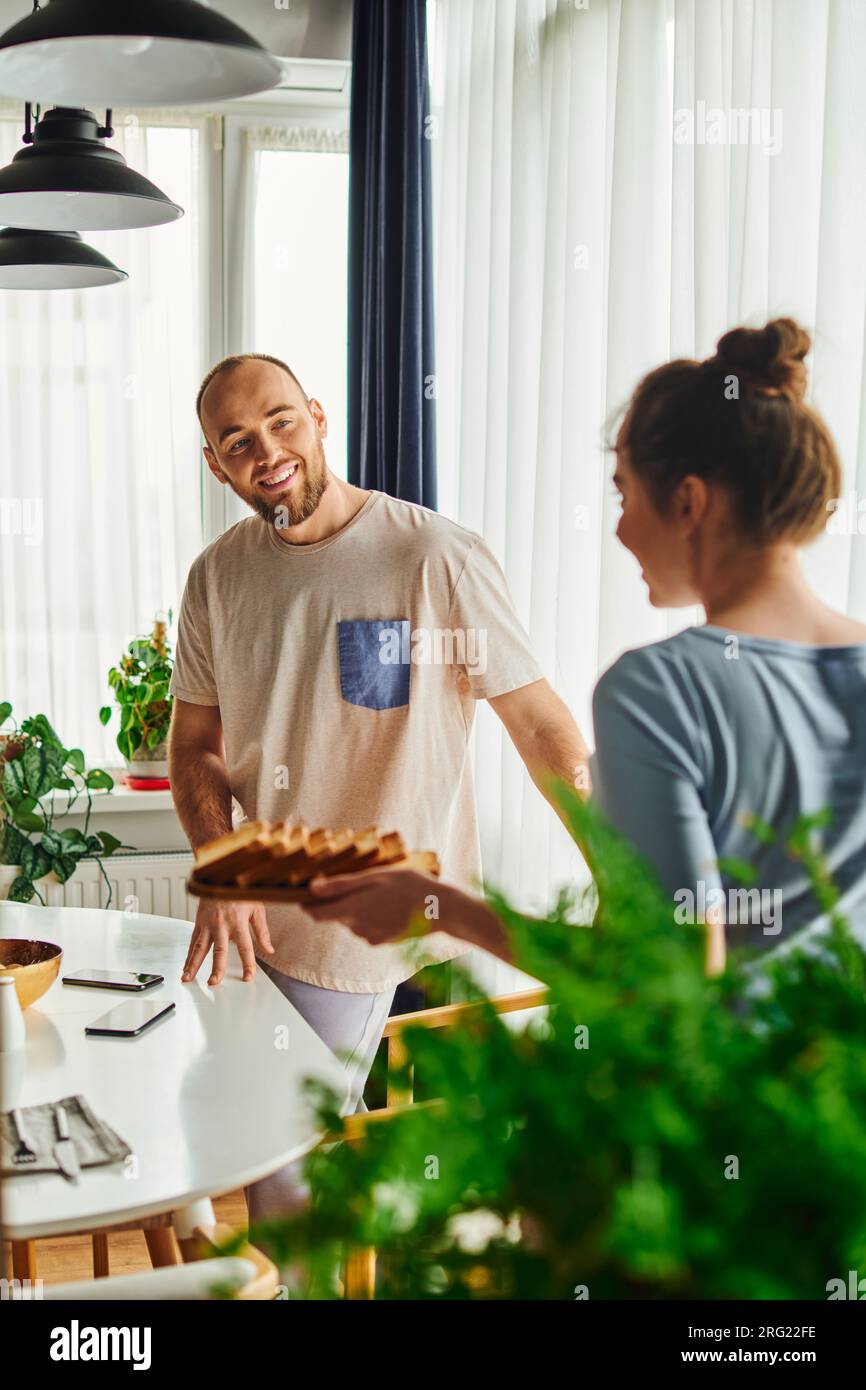 Smiling man standing near smartphones and blurred girlfriend with breakfast at home in morning Stock Photo