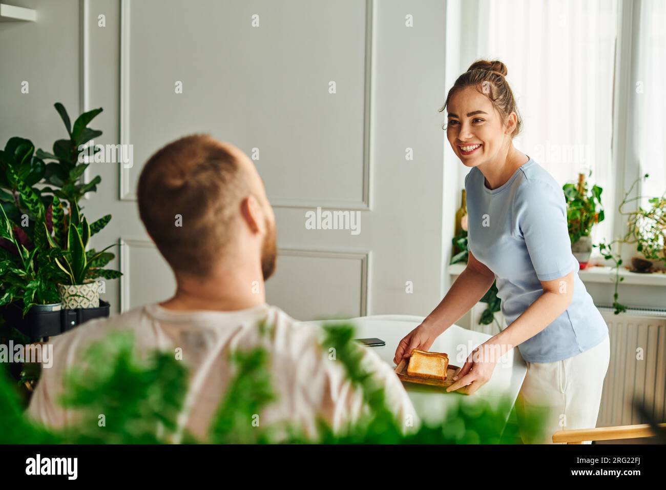 Joyful woman in homewear putting toasts on table near blurred boyfriend during breakfast at home Stock Photo