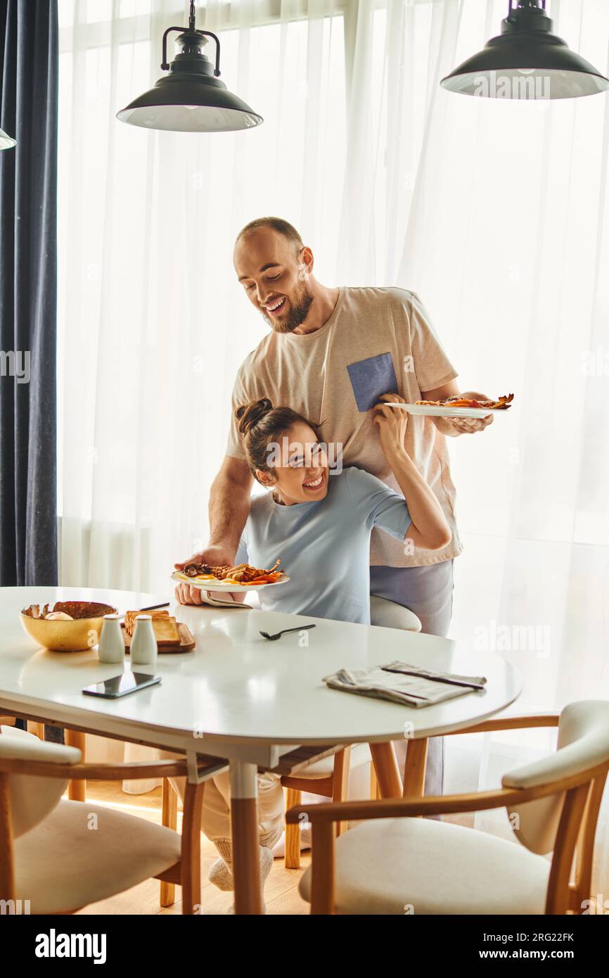 Positive woman in homewear sitting near boyfriend putting breakfast on table at home in morning Stock Photo