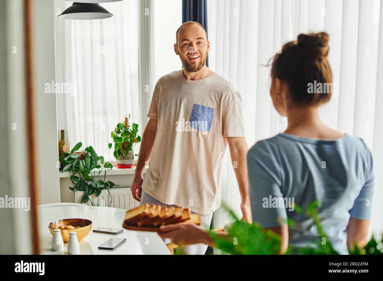 Smiling man in homewear standing near blurred girlfriend with homemade breakfast in morning at home Stock Photo