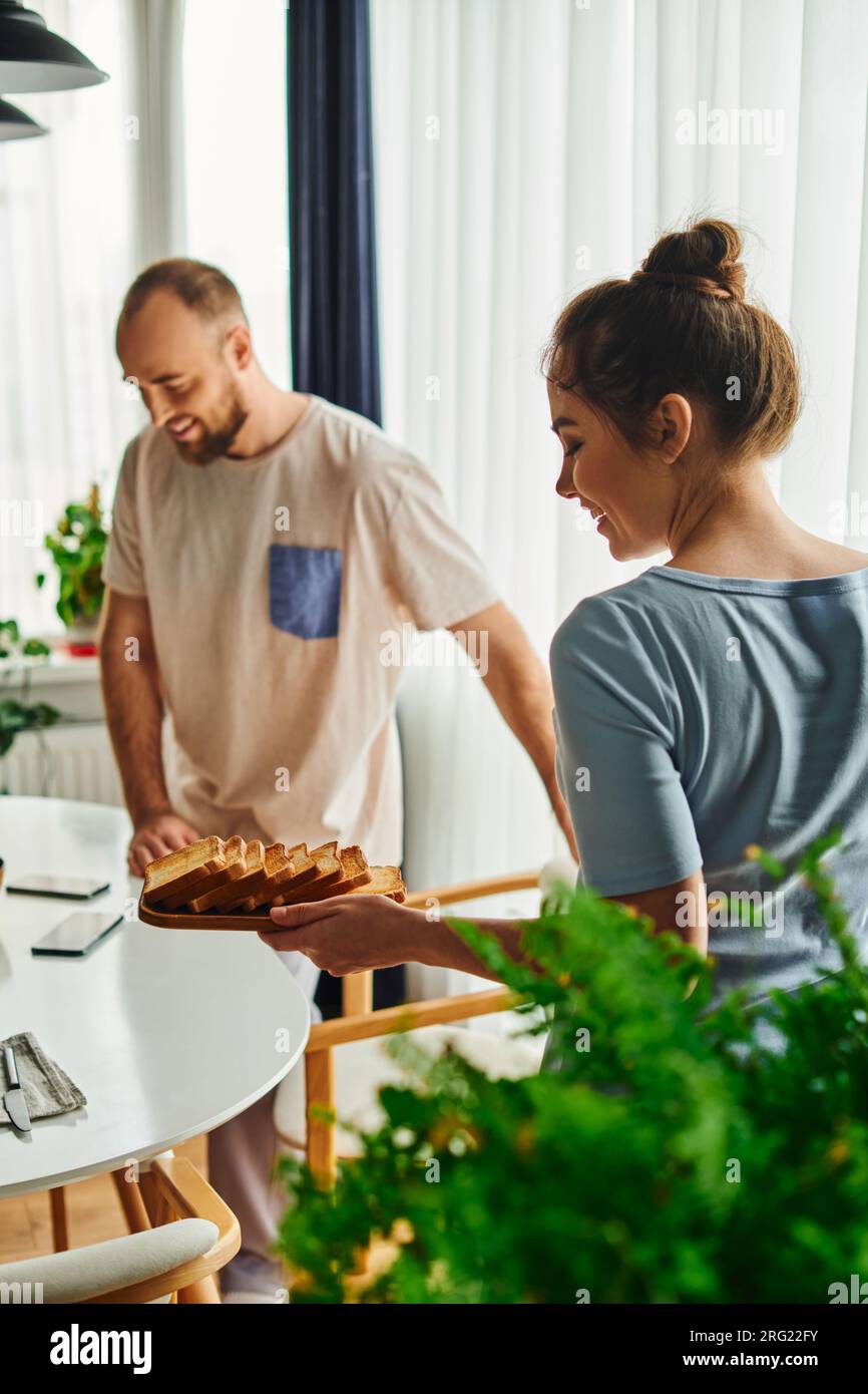 Smiling woman in homewear holding toasts near blurred boyfriend during breakfast at home in morning Stock Photo