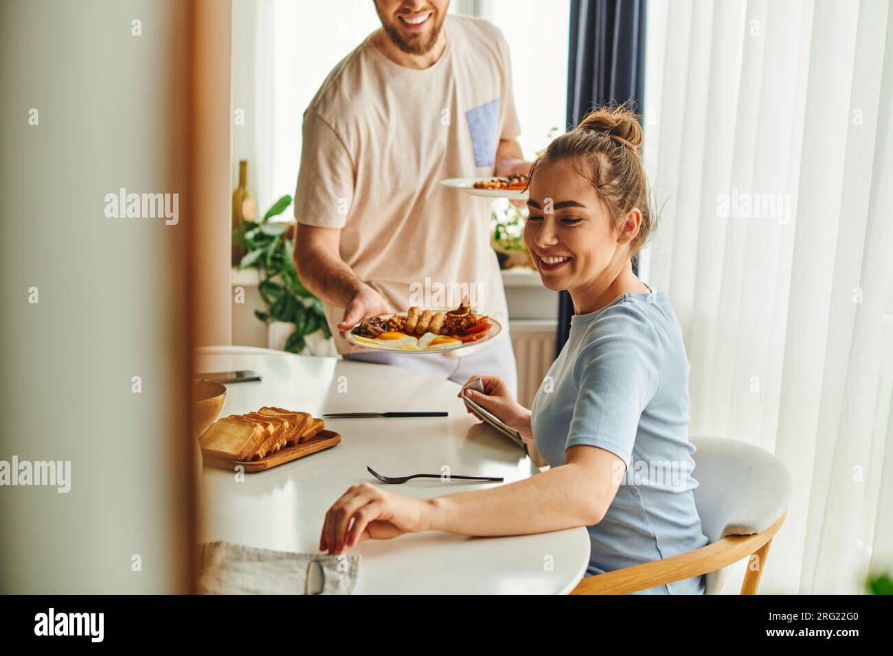 Smiling woman sitting near cutlery and blurred boyfriend holding tasty breakfast at home in morning Stock Photo