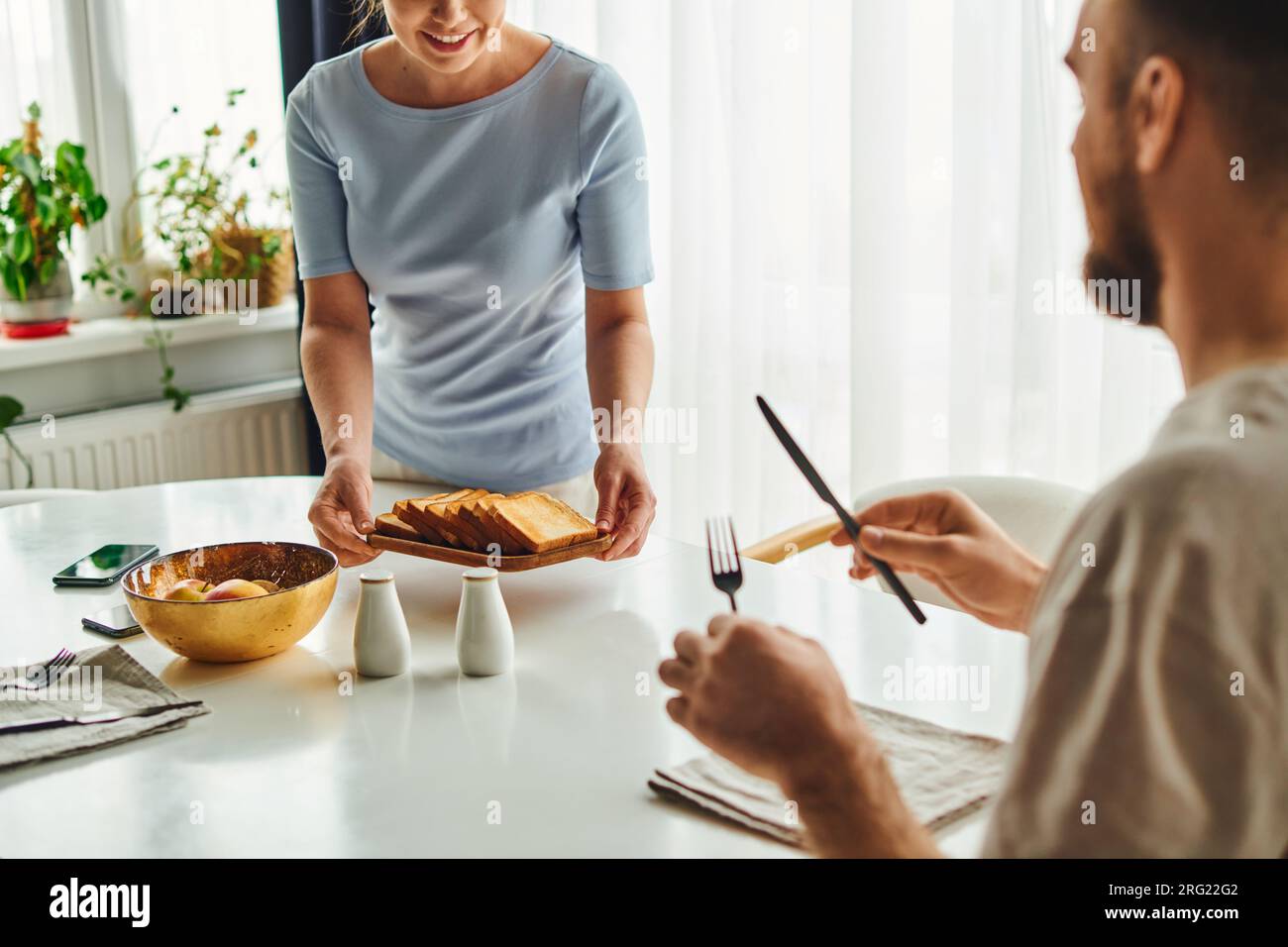 Smiling woman putting toasts on table near boyfriend with cutlery and smartphones in morning at home Stock Photo