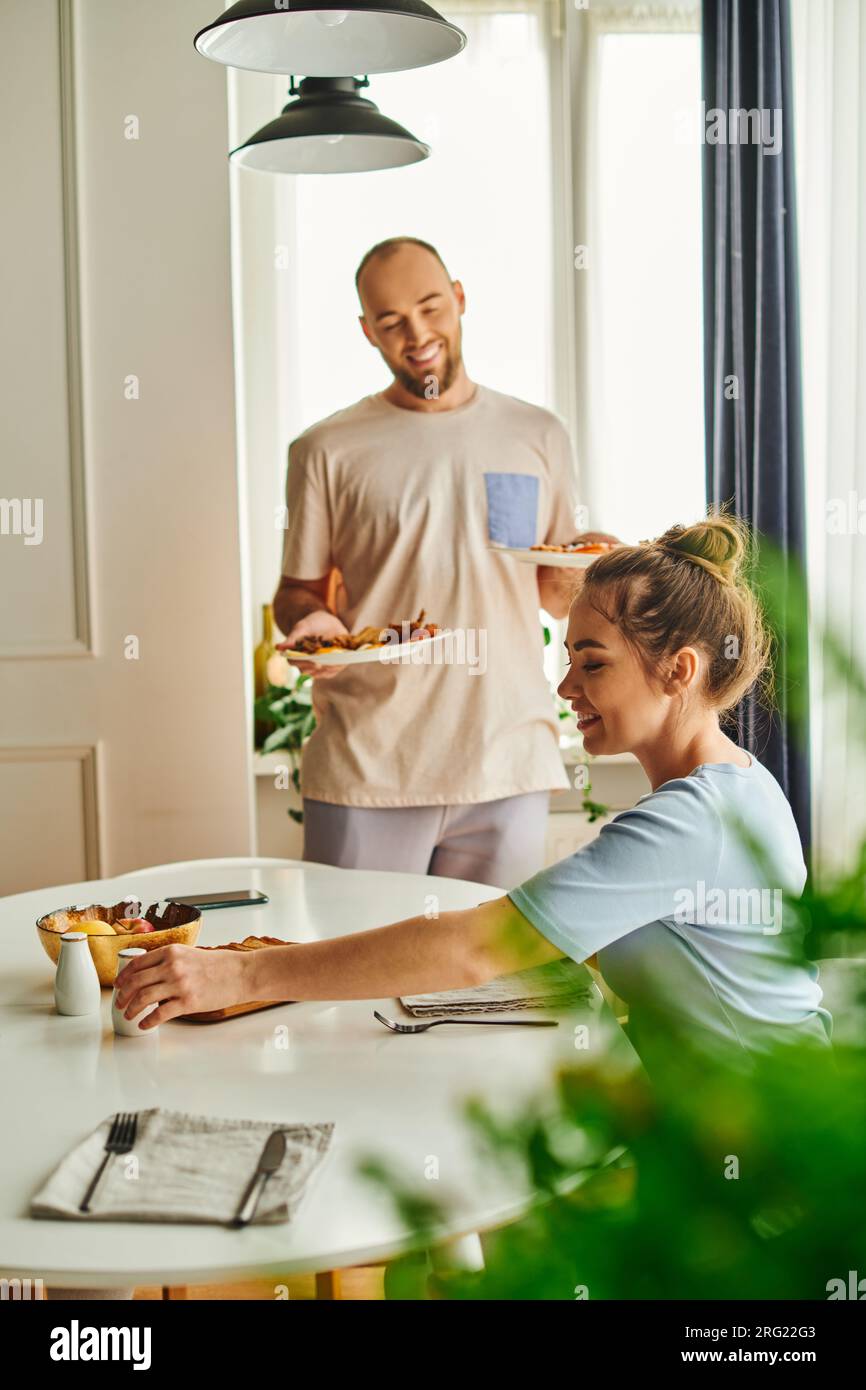 Smiling woman taking salt near blurred boyfriend with breakfast on plates at home in morning Stock Photo