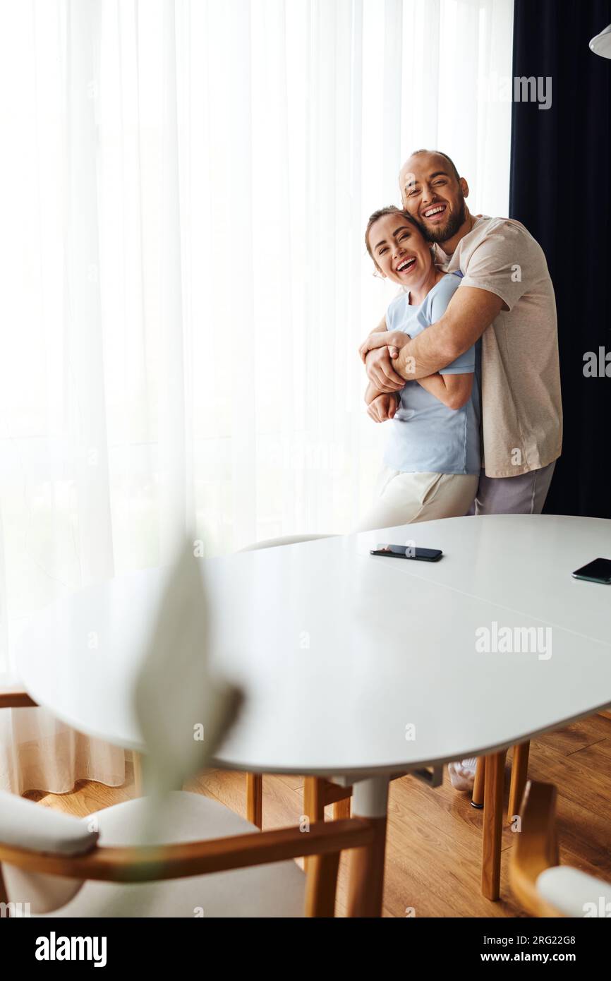 Smiling couple in homewear hugging and looking at camera near smartphones on table at home Stock Photo