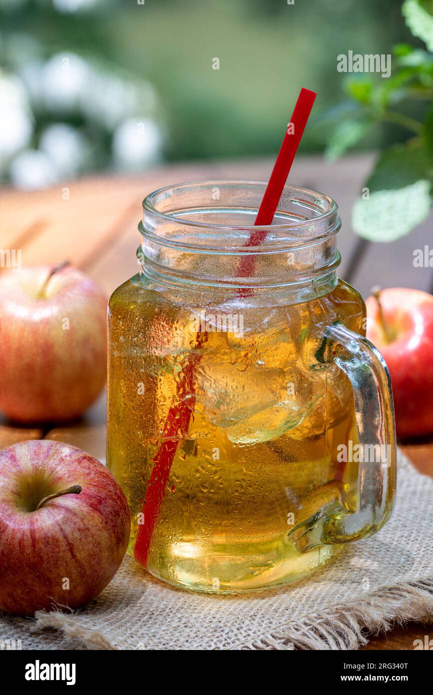 Cold apple juice in glass jar and fresh red apples outdoors on wooden patio table with nature background Stock Photo