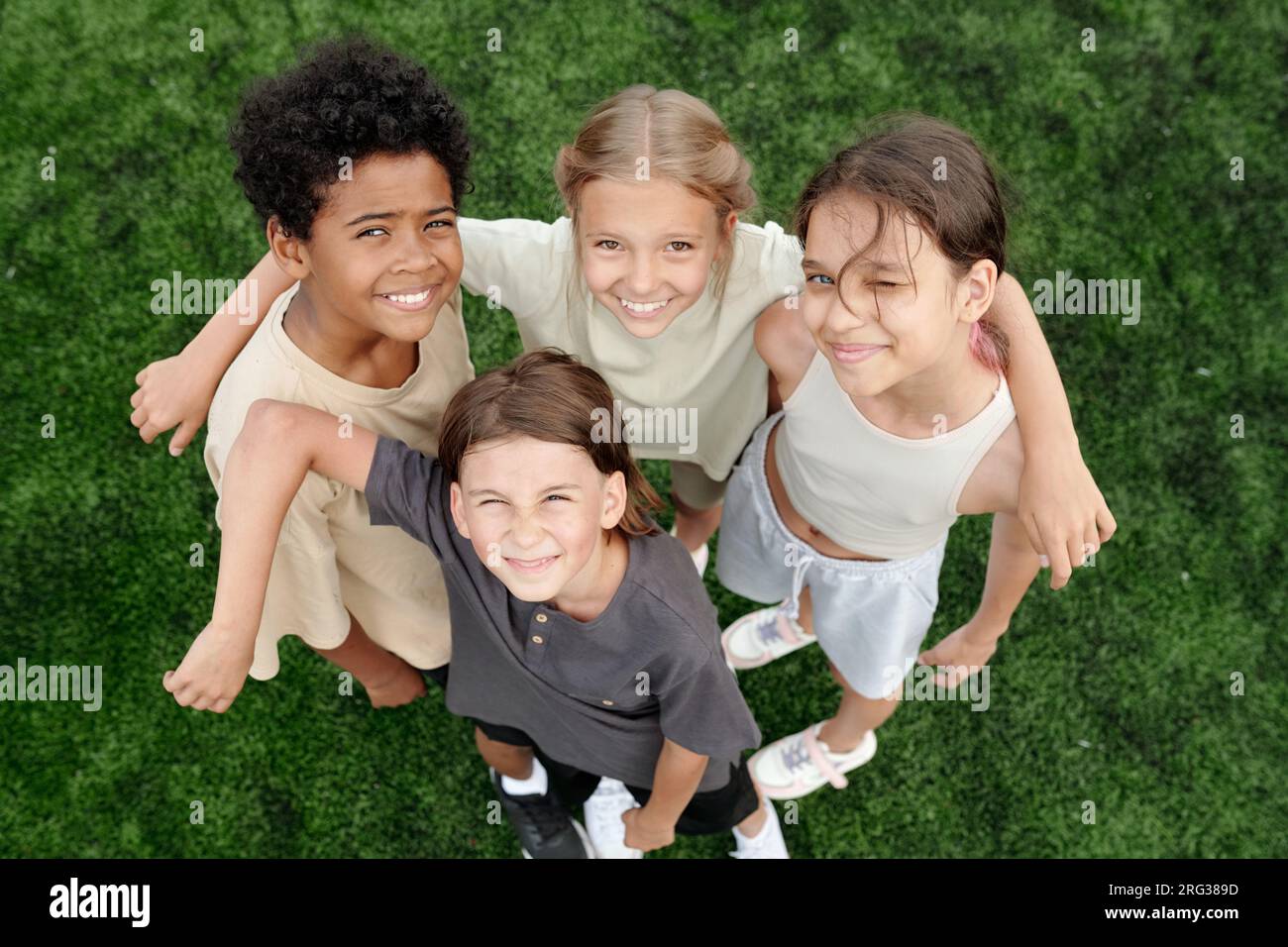 Above angle of several happy intercultural children in activewear standing against green lawn or football field and looking at camera with smiles Stock Photo