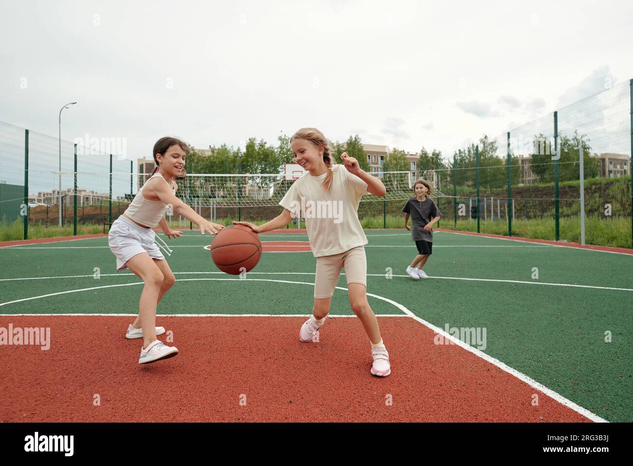 Two cute blond schoolgirls in activewear dribbling ball while running along field or stadium towards basket on background of their classmate Stock Photo