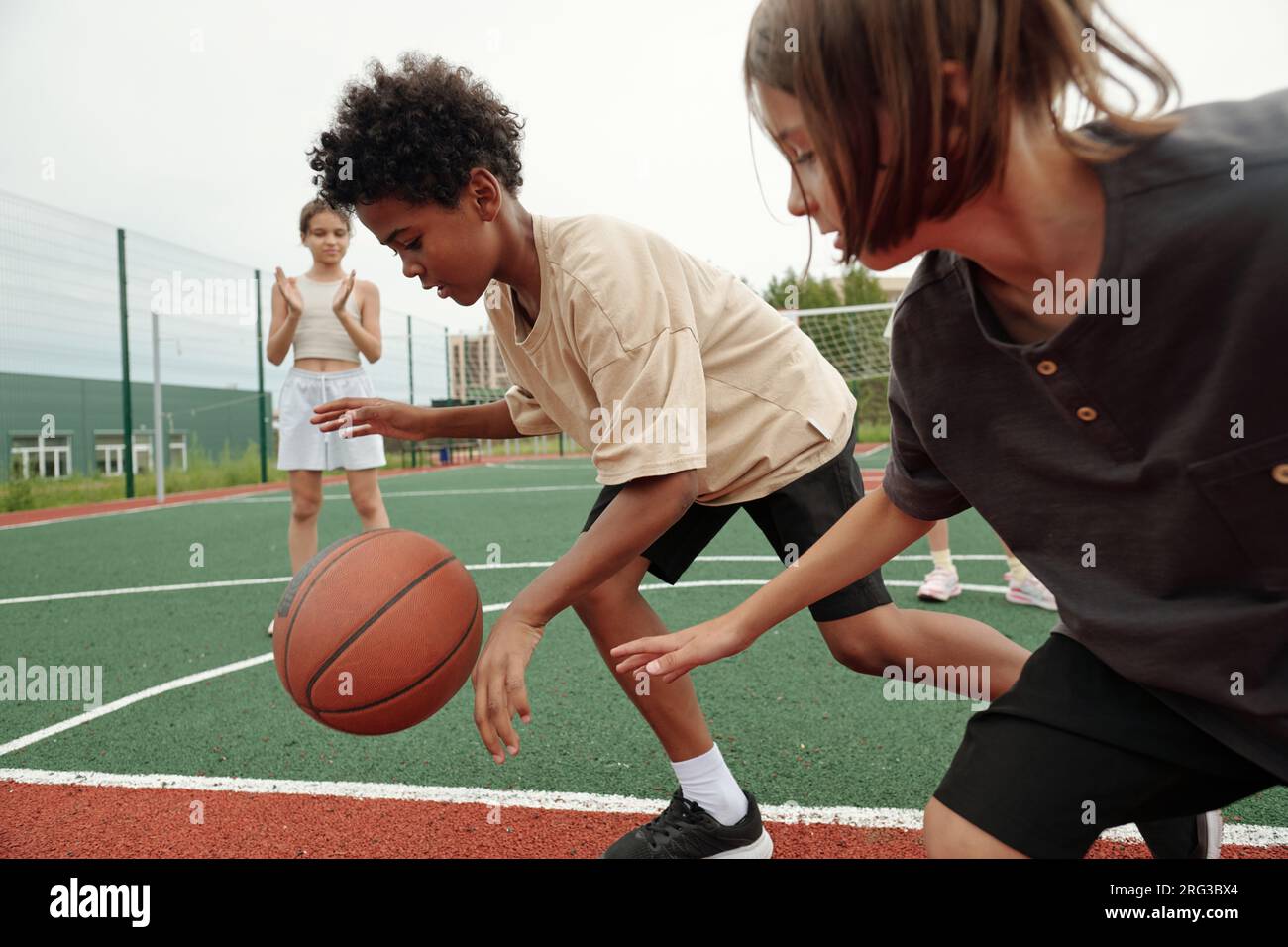 African American schoolboy in activewear dribbling ball while running forwards in front of his classmate while blond girl standing on background Stock Photo
