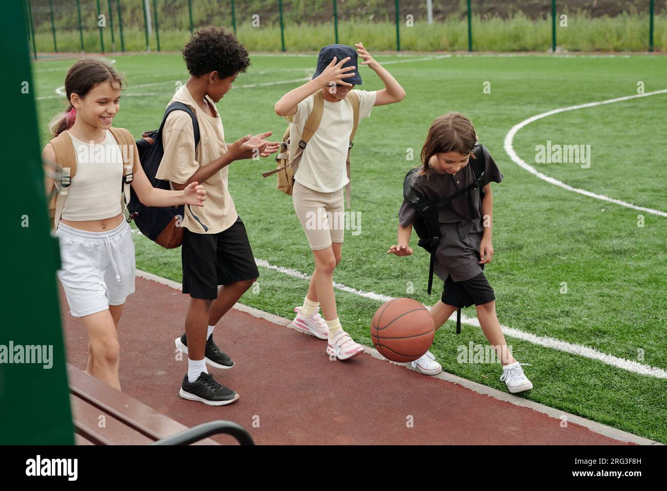 Four active children in t-shirts and shorts moving along track and green football field at modern stadium and having discussion of something Stock Photo