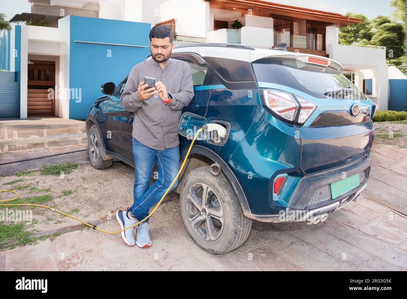 Happy Indian man using smartphone while his modern electric car getting charge outside house. Electric vehicle Recharging battery charging port. Renew Stock Photo
