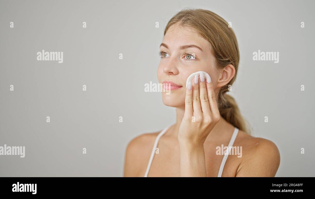 Young blonde woman cleaning face with cotton pad over isolated white ...