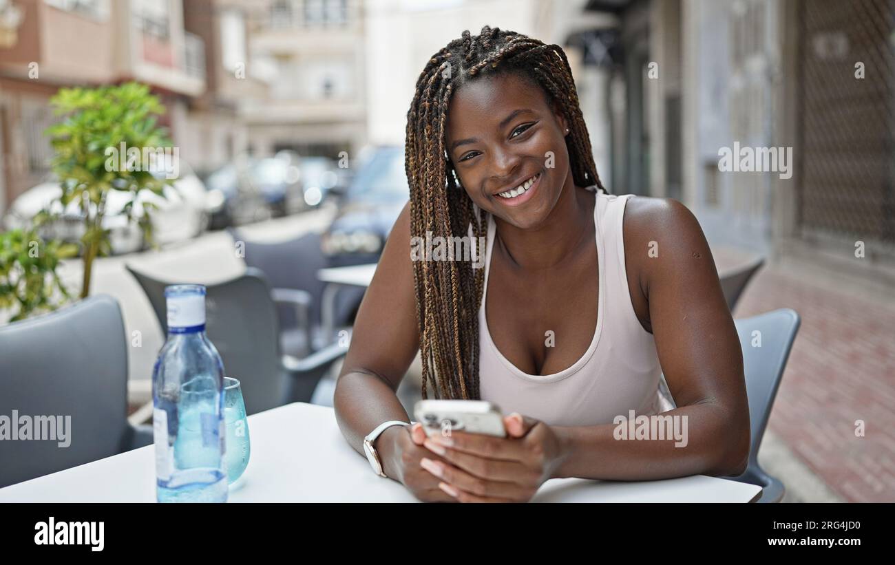 African american woman using smartphone sitting on table smiling at ...