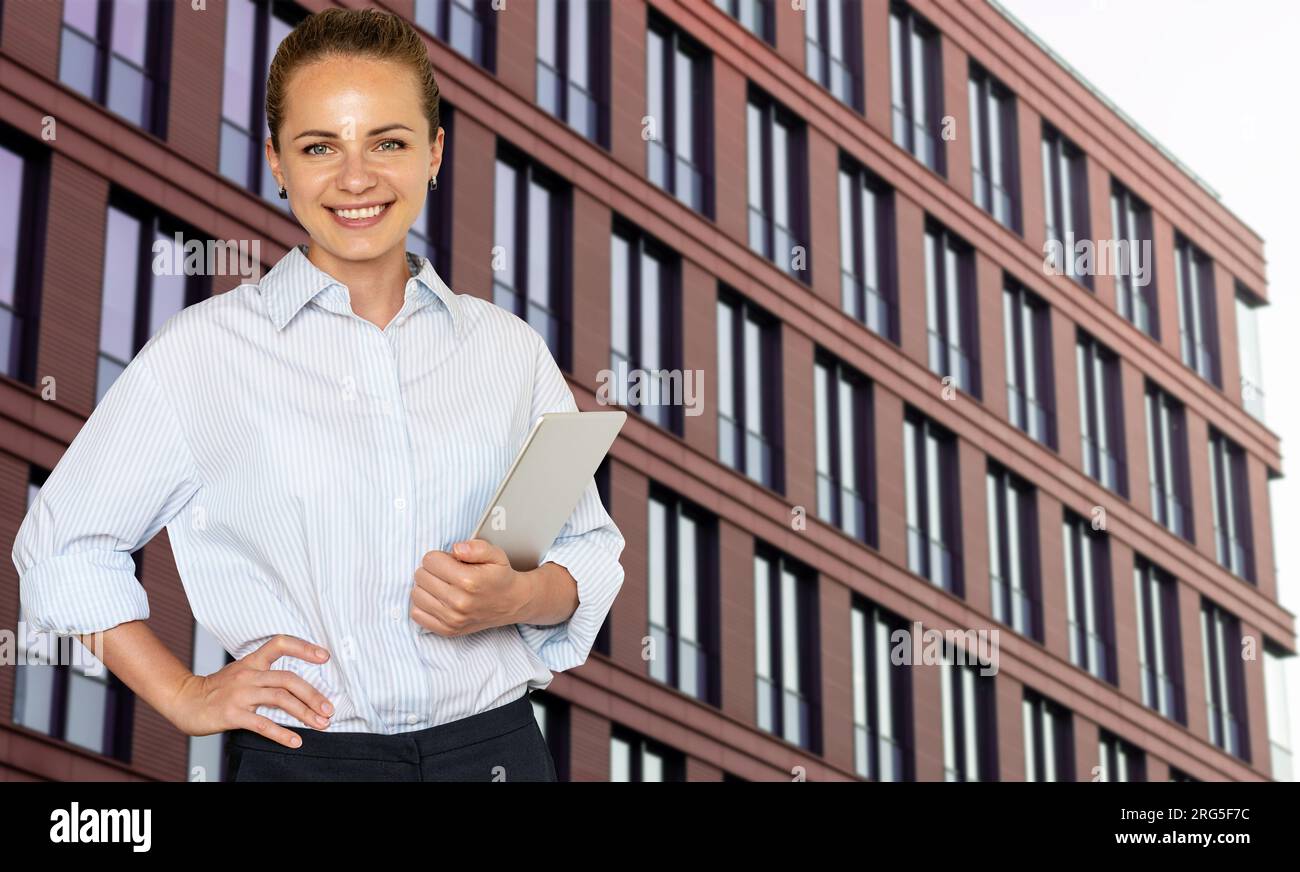 Young woman business real estate agent standing in front of business center in downtown holding digital tablet in her hand and smiling. Stock Photo