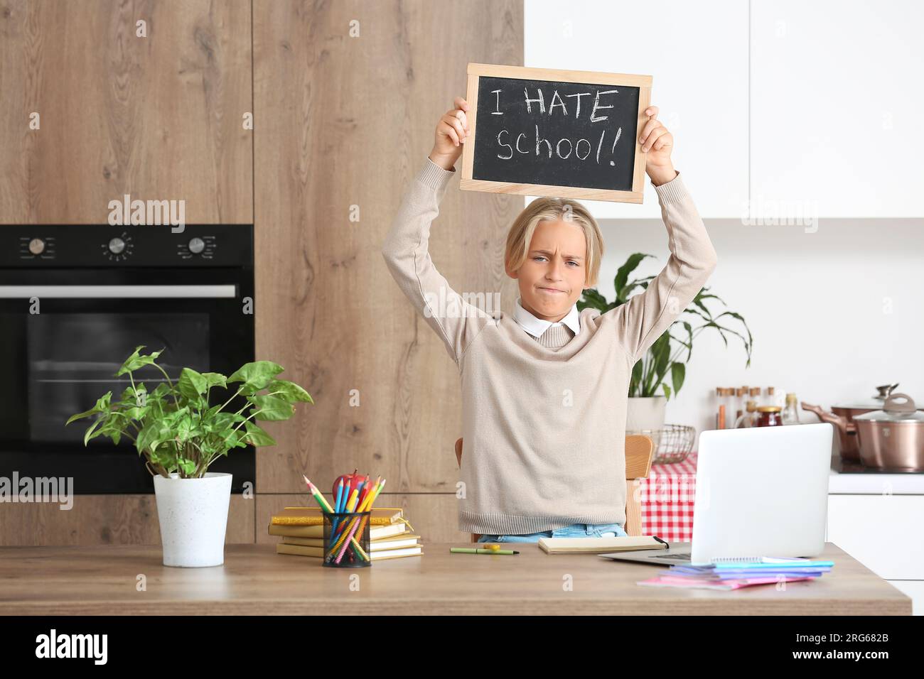 Displeased little boy holding chalkboard with text I HATE SCHOOL in kitchen Stock Photo