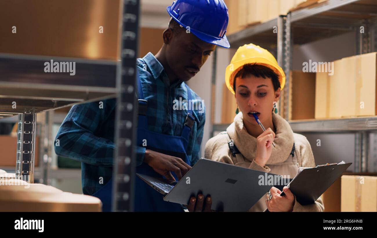 Multiethnic team of workers analyzing list on laptop and tablet, working with industrial cardboard boxes in storage room. People checking merchandise stock for delivery service. Handheld shot. Stock Photo