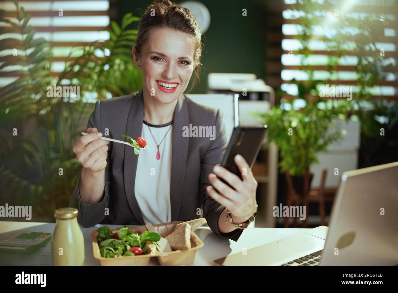Sustainable workplace. smiling modern 40 years old woman worker in a grey business suit in modern green office with laptop and smartphone eating salad Stock Photo