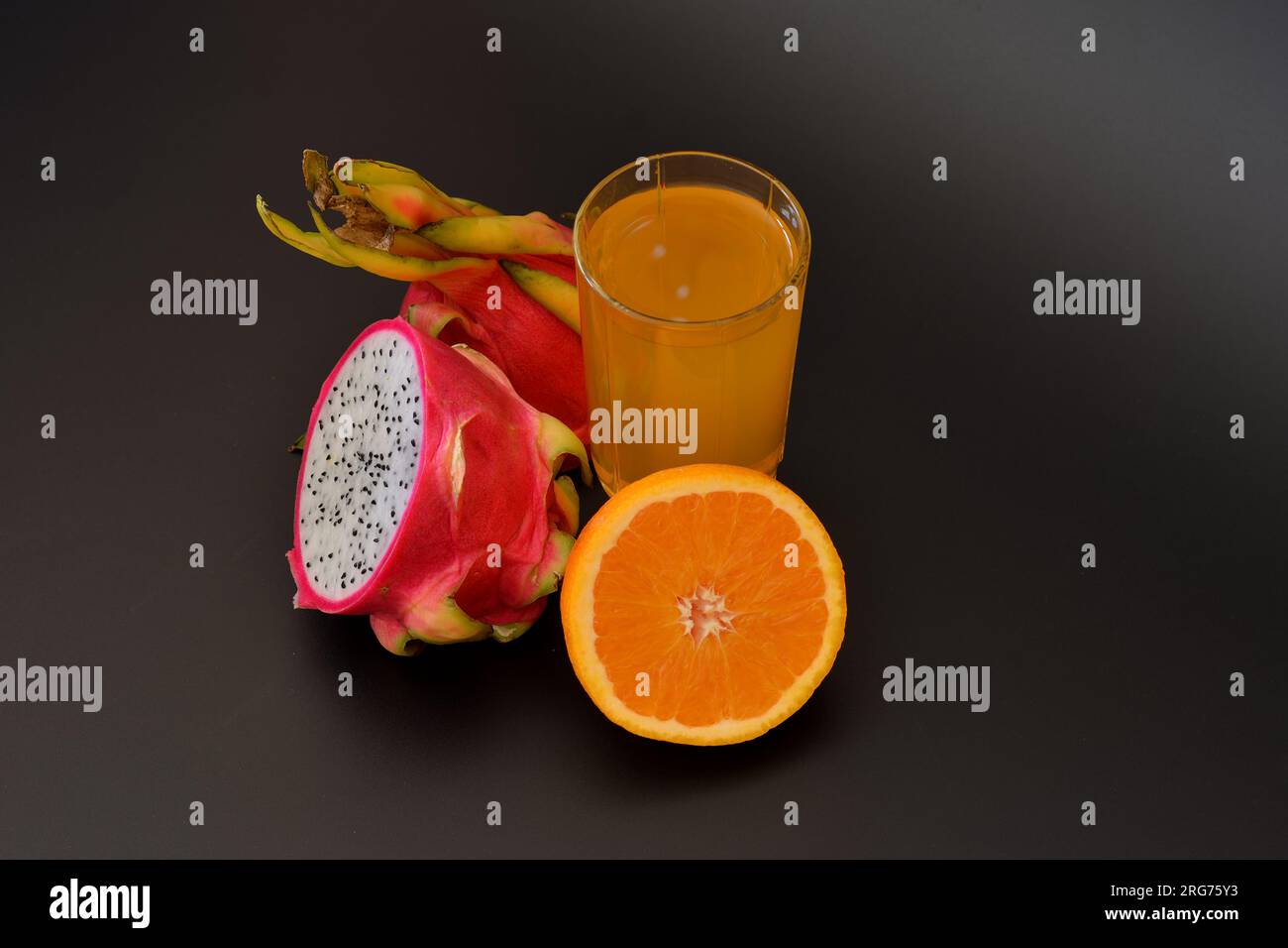 A tall glass of fruit juice on a black background, next to ripe slices of pitaya and orange. Close-up. Stock Photo