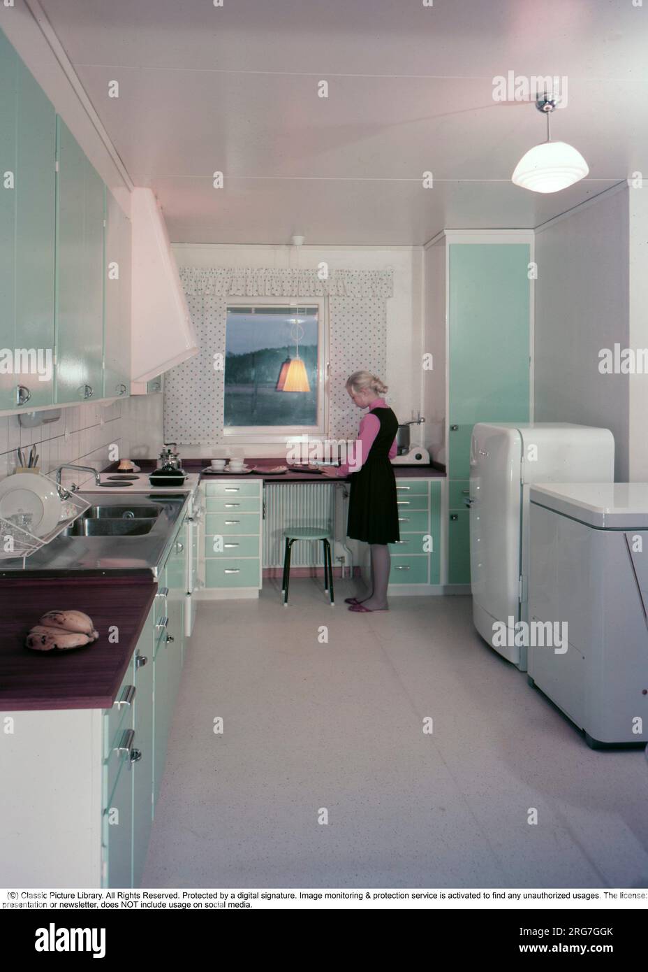 In the early 1960s. A lovely color picture of a typical 1950s - 1960s kitchen with the cupboards and kitchen doors painted in a green color. It is the decade of the first well planned and organized kitchen and it's functions to work in and have room for the appliances like the electric cooker, the freezer and the refrigirator. She is standing by the kitchen bench. Sweden 1960. Conard ref BV45-3. Stock Photo