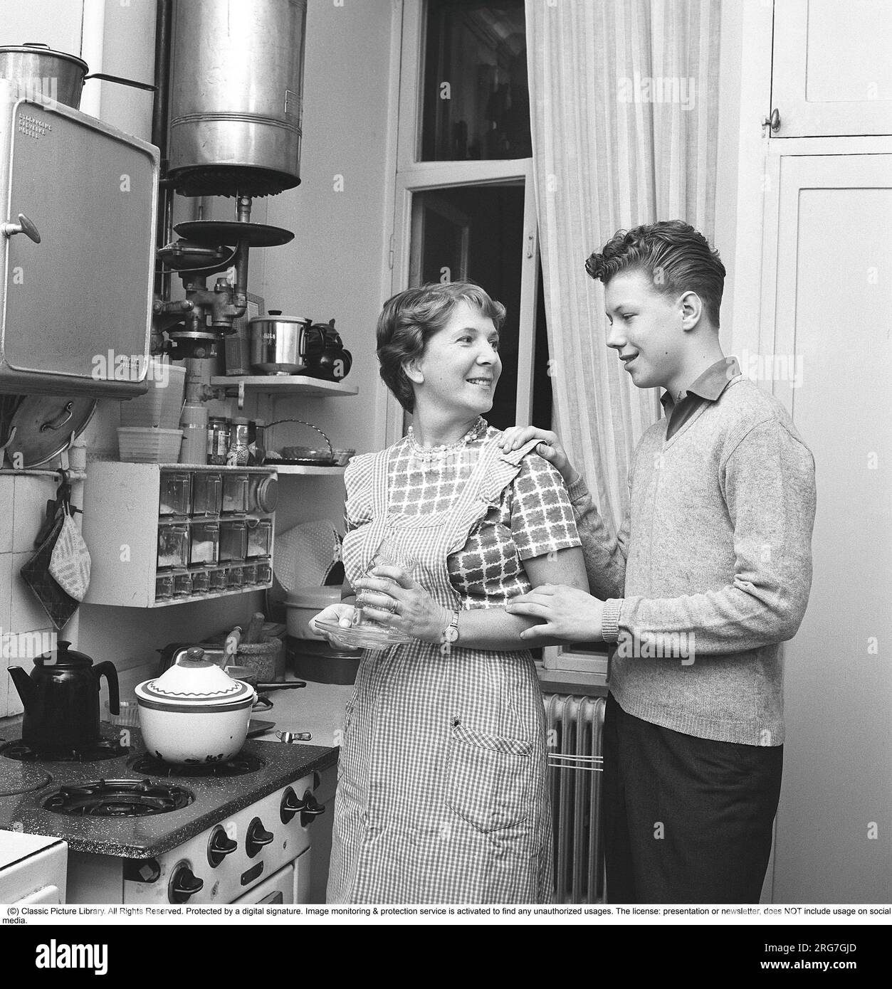 Kitchen in the 1960s. A mother with her son in the kitchen. A small kitchen with a gas stove, a smaller kitchen bench where there is a mortar and bread box. A spice rack hangs on the wall. A water heater hangs at the top of the wall. Sweden 1962. Kristoffersson ref CV90-10 Stock Photo
