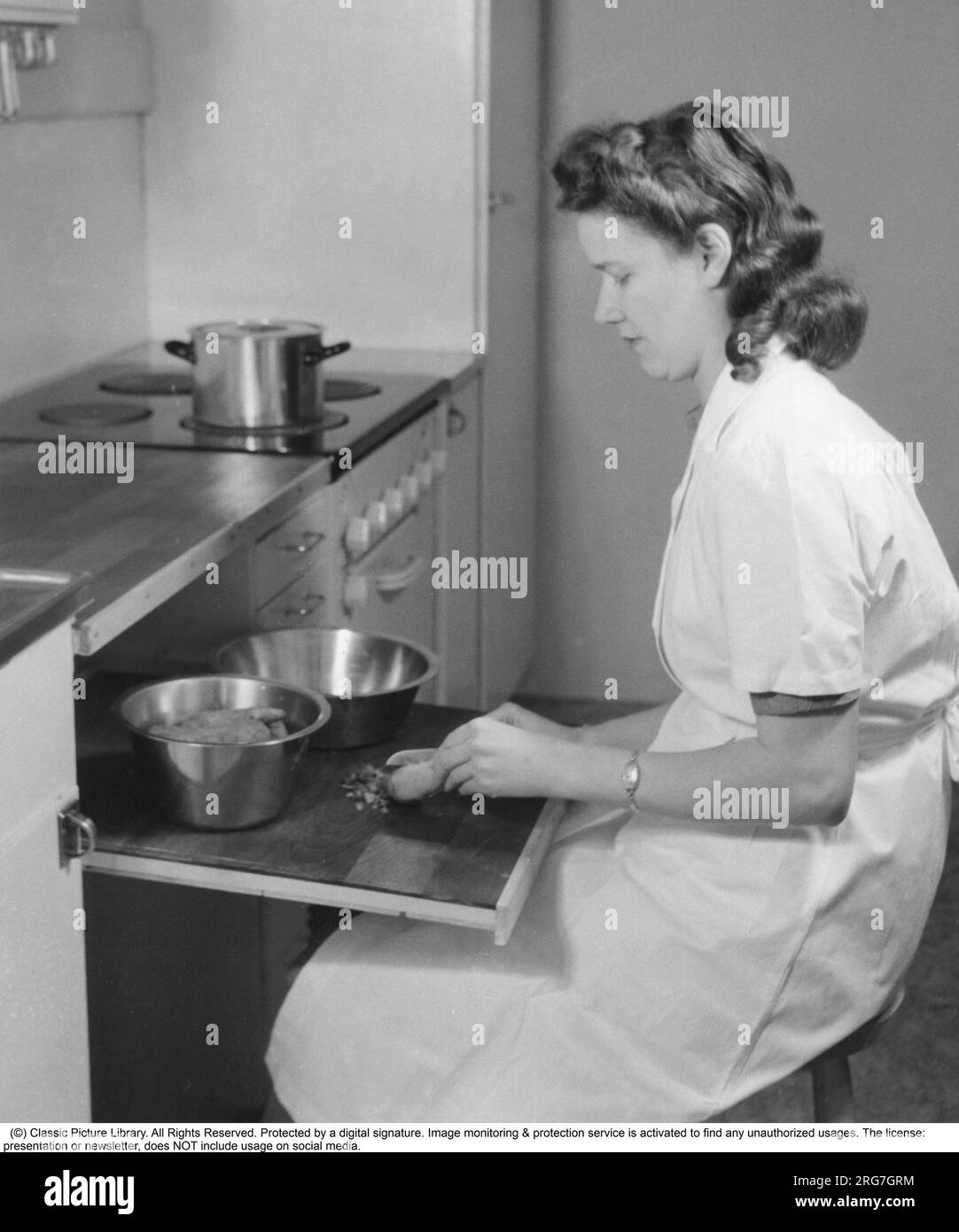In the 1940s. An woman is seen sitting at the practical extendable cutting board, preparing the ingredients for a a dinner meal. Sweden 1944. Stock Photo