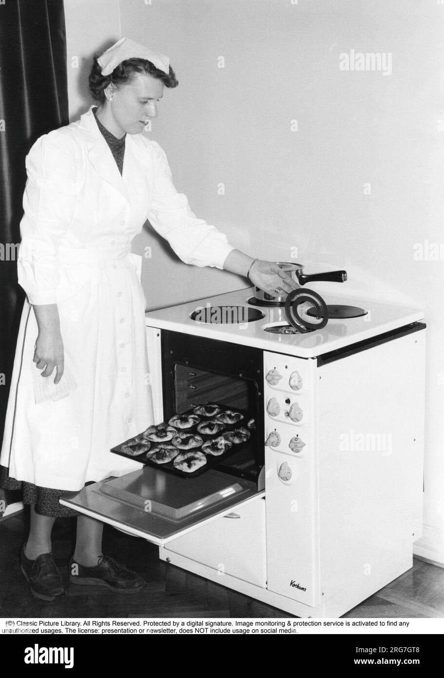 In the kitchen in the 1950s. A young woman in the kitchen at the brand new Kockums electric cooker. She is seen demonstrating the practical function of lifting the hotplate in order to clean properly under and around it. A plate of freshly baked pastries is visible. Sweden 1957 Stock Photo