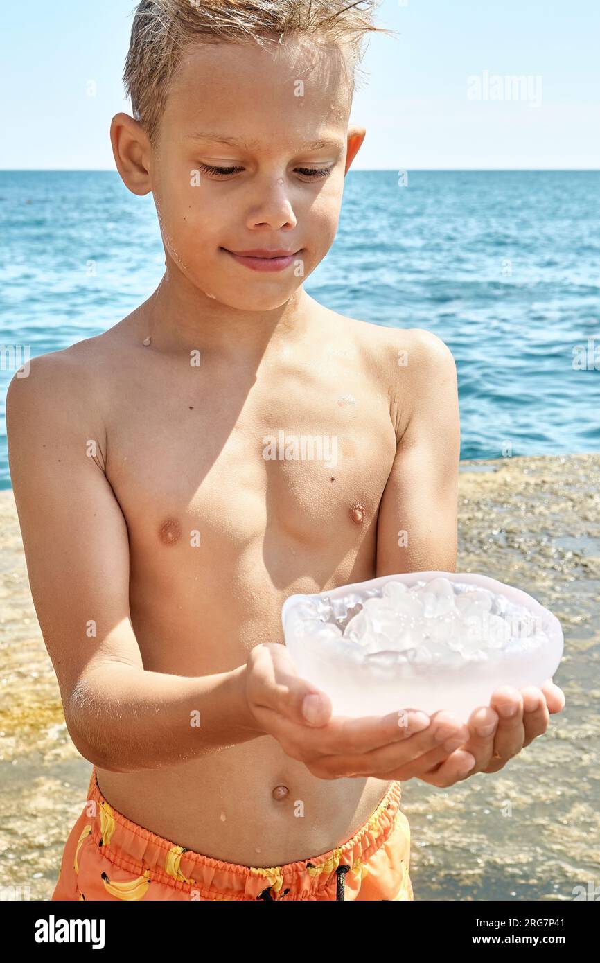 Boy standing on stone pier holds transparent jellyfish with amused expression looks at jellyfish in hands Stock Photo