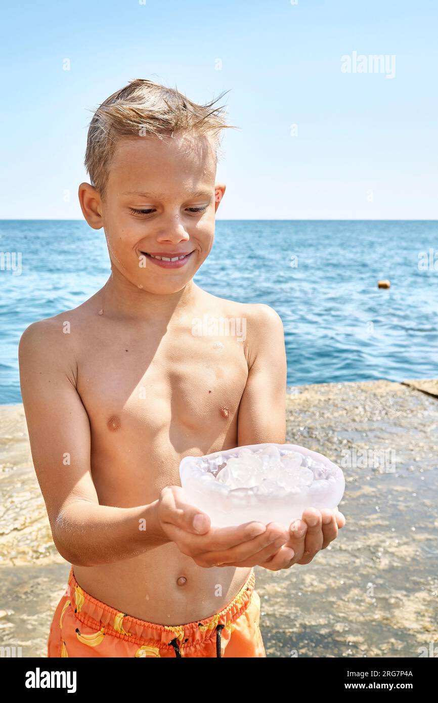 Boy standing on stone pier holds transparent jellyfish with amused expression looks at jellyfish in hands Stock Photo