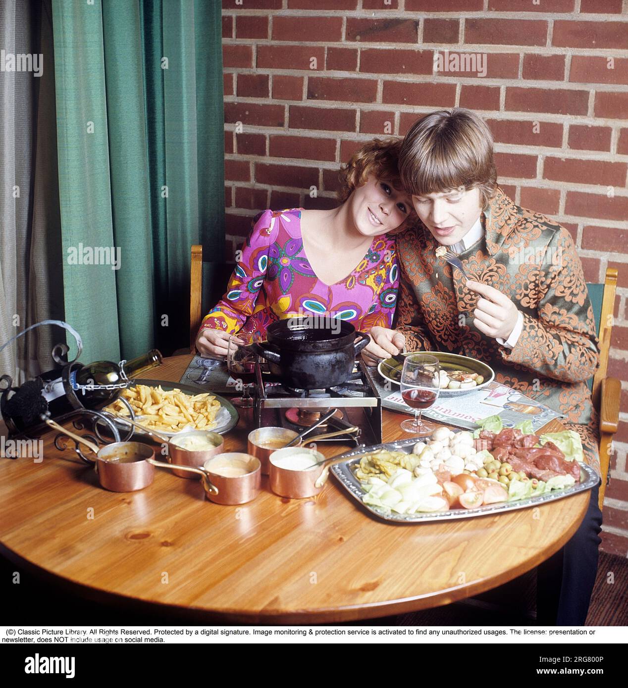 Fondue. A dish that had its breakthrough in the 60s and 70s. A fondue pot with hot oil stands in the middle of the table and you dip meat into it to cook and eat yourself. She is wearing a very colorful and patterned long sleeved blouse that was typical of the late 60's and 70's. Him in a pop inspired jacket not unlike the clothing that singers wore at the time. Sweden 1970. Conard ref CV49-5 Stock Photo