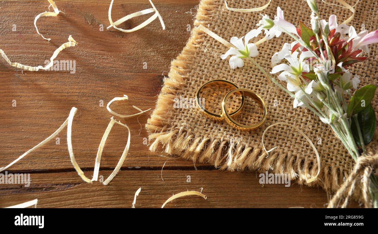 Detail of two gold rings on sackcloth on a rustic wooden table with straw and a bouquet of flowers. Top view. Stock Photo