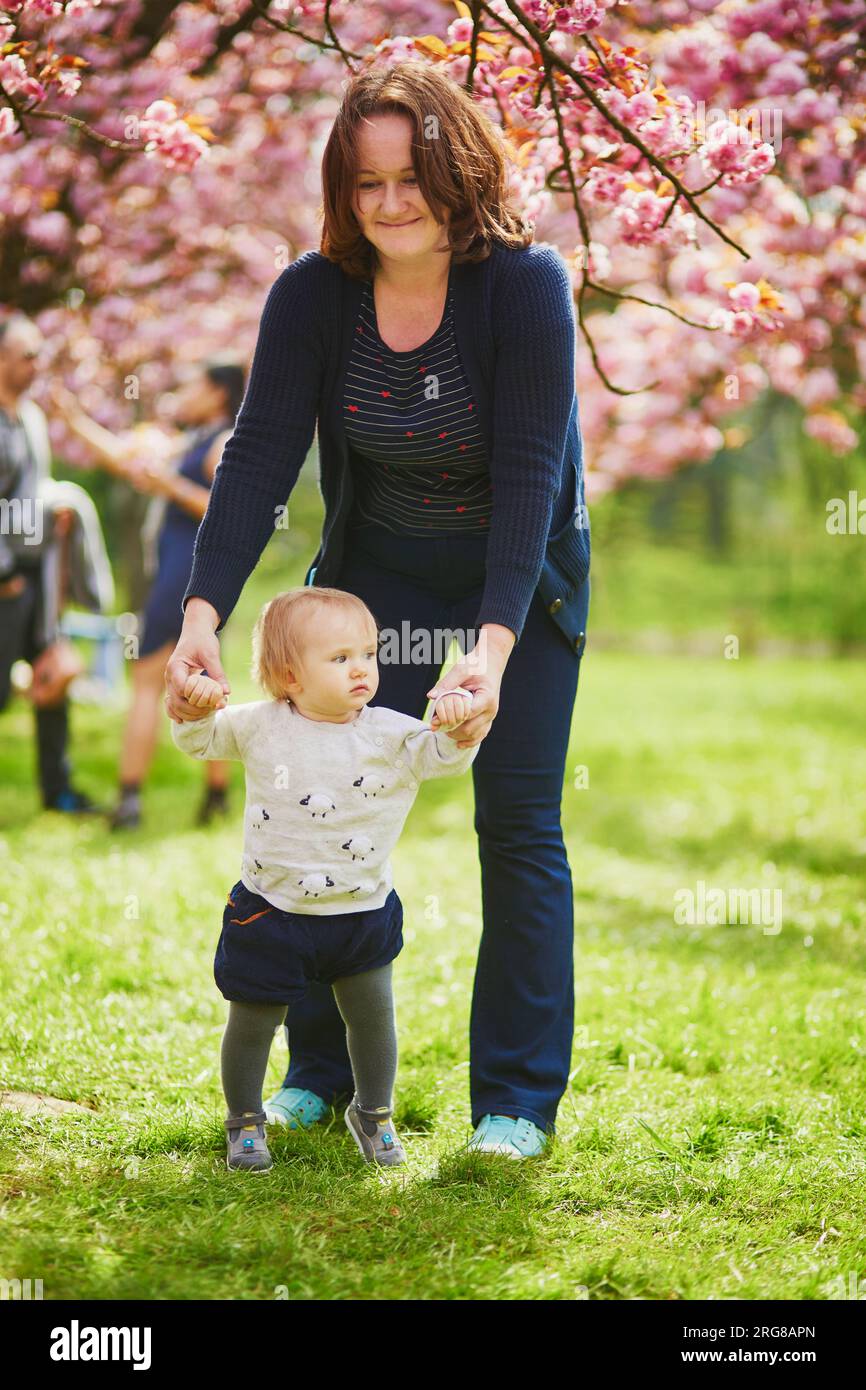 Young woman with 1 year old baby girl in park on a spring day, enjoying cherry blossom season. Mother helping her daughter make first steps. Toddler l Stock Photo