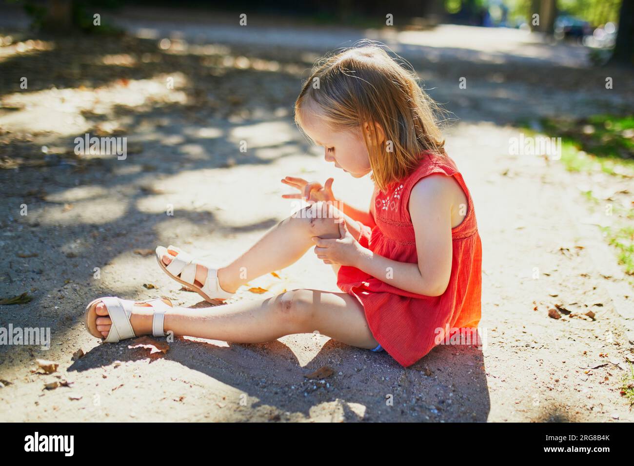 Cute little girl sitting on the ground after falling down. Child getting hurt while walking in park. Kid looking at her boo-boo Stock Photo
